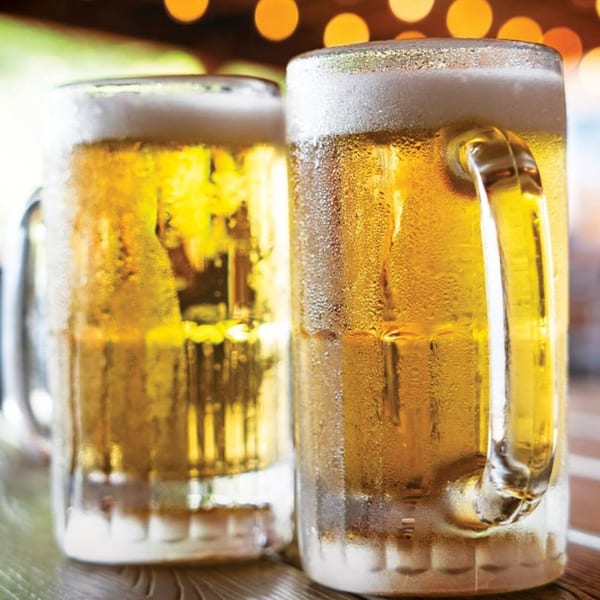 Two frosty mugs of golden beer with foamy heads sit on a wooden table with bokeh lights.