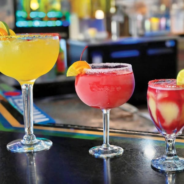 Three colorful margaritas, yellow, red, and swirl, garnished with fruit on a bar.