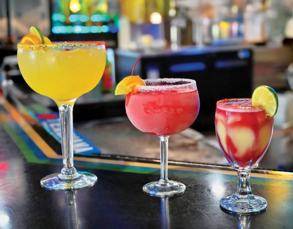 Three colorful margaritas, yellow, red, and swirl, garnished with fruit on a bar.