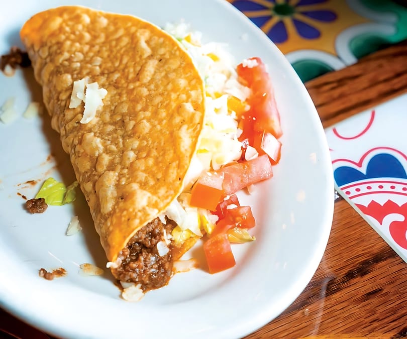 A crispy taco filled with ground beef, lettuce, cheese, and diced tomatoes on a white plate.