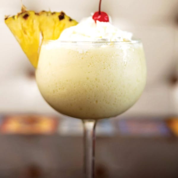 Creamy yellow cocktail in a stemmed glass, garnished with a pineapple slice, whipped cream, and cherry.