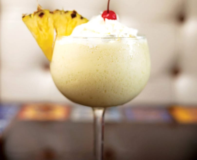 Creamy yellow cocktail in a stemmed glass, garnished with a pineapple slice, whipped cream, and cherry.