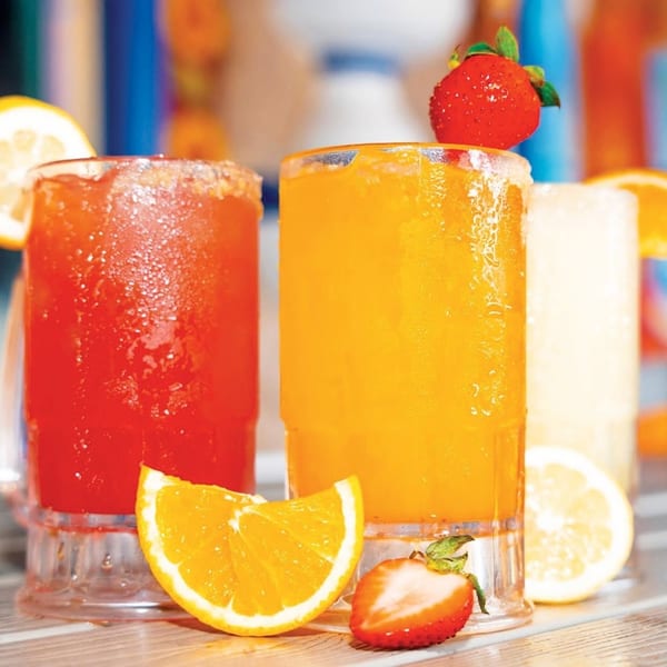 Three colorful frozen drinks, garnished with lemon, orange, and strawberries, sit on a wooden surface.
