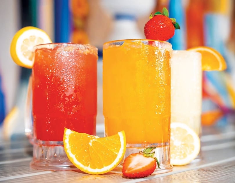 Three colorful frozen drinks, garnished with lemon, orange, and strawberries, sit on a wooden surface.