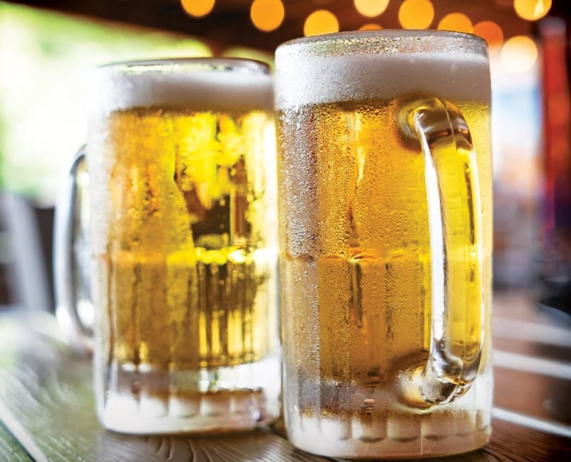 Two frosty mugs of golden beer with foamy heads sit on a wooden table with bokeh lights.